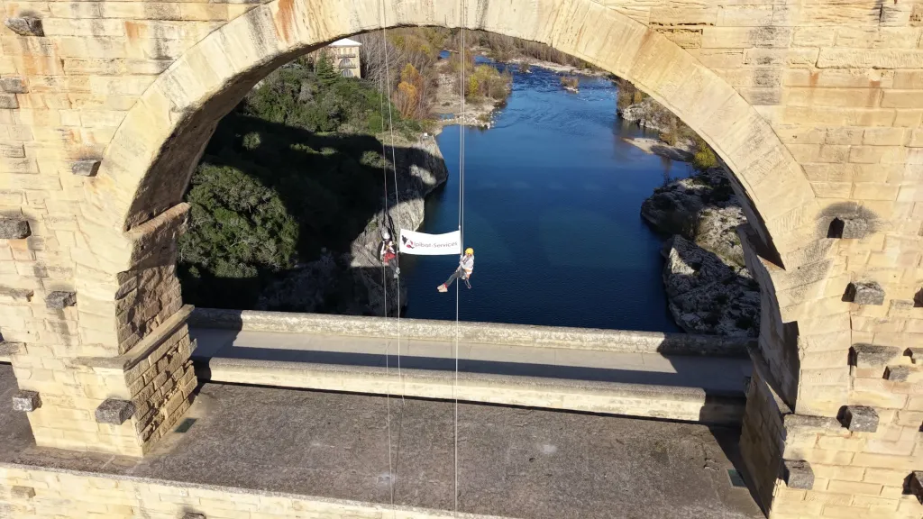 Deux cordistes d’Alpibat-Services suspendus sous une arche du pont du Gard, au-dessus du Gardon, avec une banderole de l’entreprise